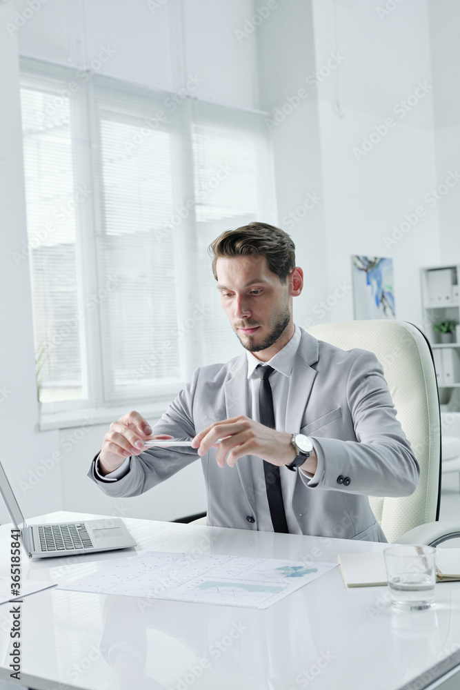 Poster handsome young manager in gray suit sitting at desk and photographing graphs on smartphone
