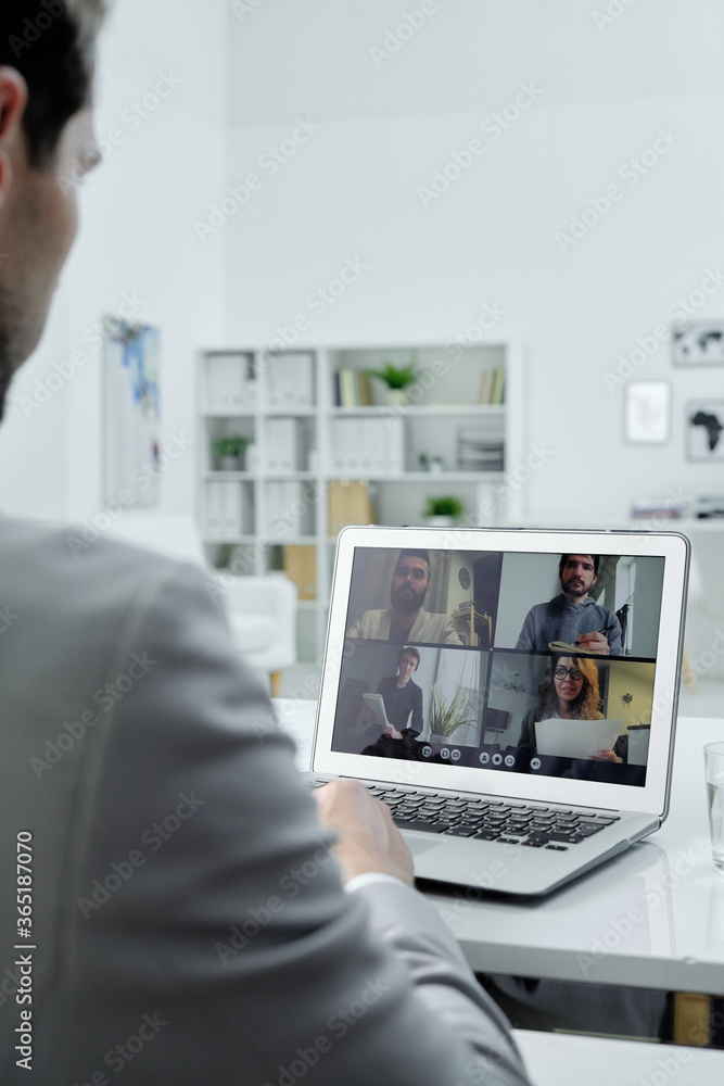 Poster over shoulder view of businessman sitting at desk in office and talking to colleagues via video conf