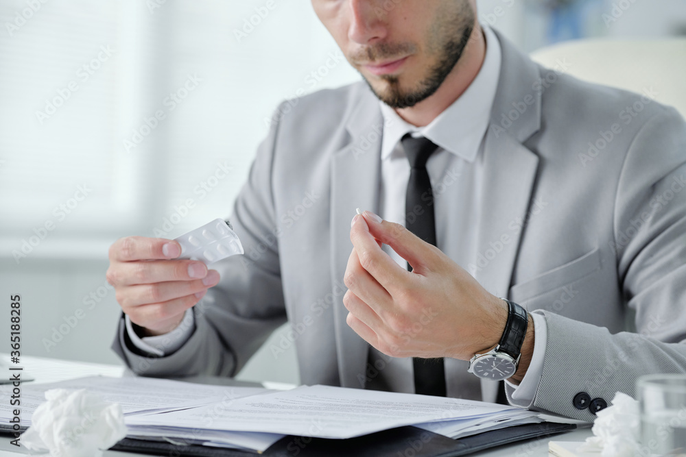 Poster Close-up of bearded businessman in gray suit sitting at table with documents and taking pill to prevent cold