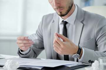 Close-up of bearded businessman in gray suit sitting at table with documents and taking pill to prevent cold