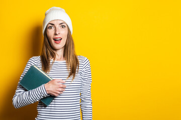 Surprised young woman in a hat holds a book on a yellow background.