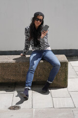 trendy girl with sunglasses, sitting on concrete bench and using smartphone