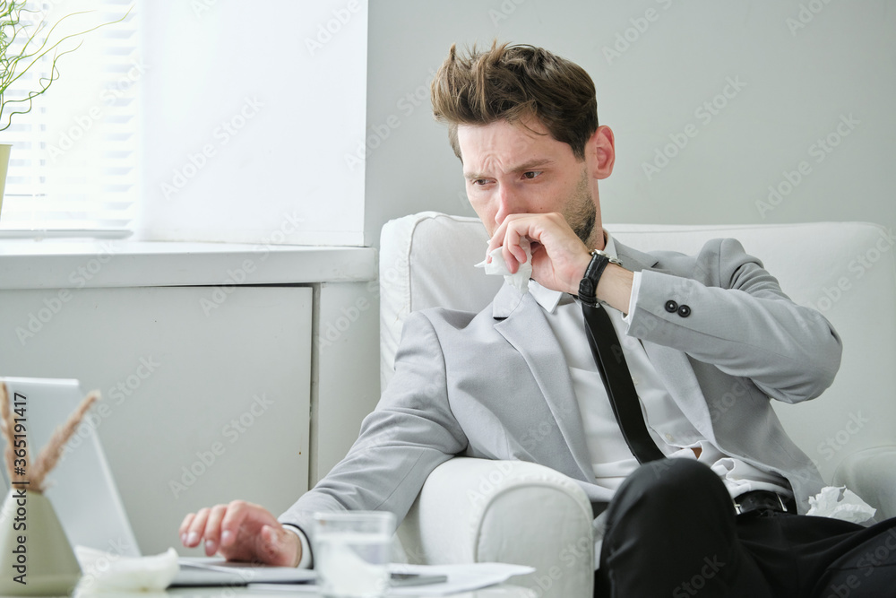 Poster frowning ill young businessman sitting in armchair and working on project using laptop while having 