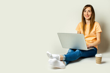 Naklejka premium Smiling young woman holding a laptop on her knees, a paper cup with coffee is standing nearby on a light background