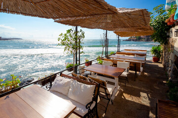 empty terrace of a restaurant on the sea. beautiful view in to the bay in morning light. decorative umbrella above wooden tables and seats. sunny weather