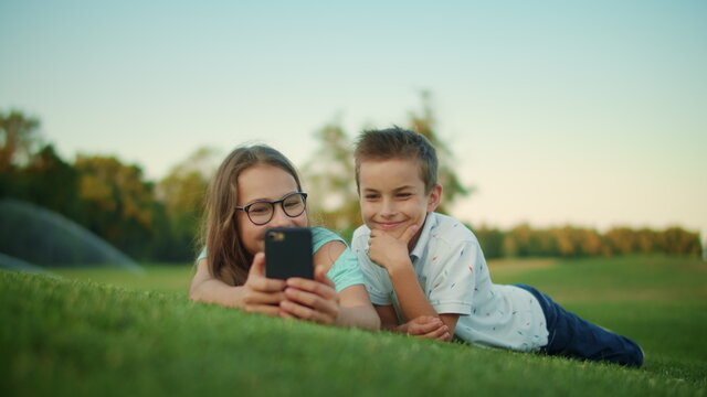 Siblings lying on grass in field. Laughing girl and boy using smartphone outdoor