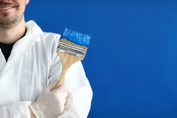 Close-up of paintbrush covered with blue bright paint. Smiling male worker in uniform posing on wall background. Professional interior designer. Copy space in right side. Construction site concept
