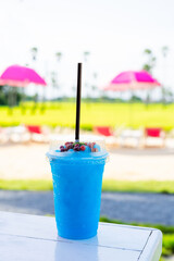 Blue Hawaii smoothie drink topped with berries Put on a white wooden table The background is a purple seaside seat with a view of natural green fields.