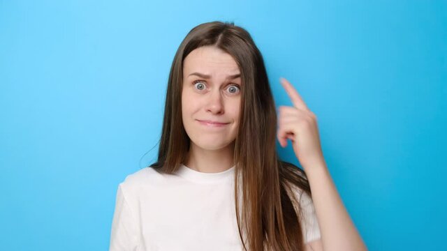 Annoyed young woman pointing at camera and showing stupid gesture, blaming some idiot, dressed in white t-shirt, isolated on blue studio background. Portrait of brunette girl in despair and shock