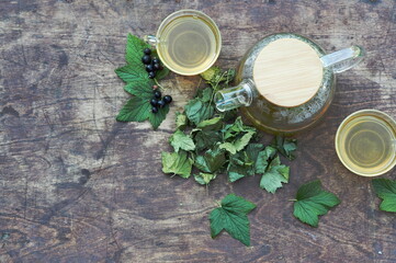 Tea in a glass teapot with cups with green leaves of black currant on a wooden colorful table.