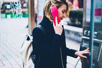 Young female traveler using old fashioned public telephone for making international calls on trip, pensive hipster girl dialing number on metal payphone operated with coins for having conversation