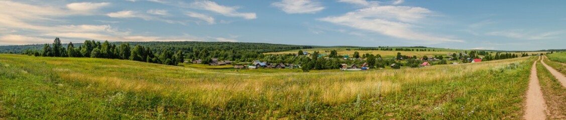 Panoramic view of a small village, in the foreground a field with a dirt road, in the background a forest. Udmurt Republic, Russia.