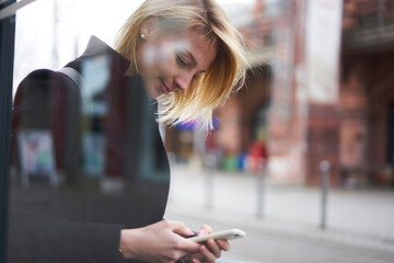 Attractive blonde female traveler dialing number of taxi standing on blurred urban setting copy space area for advertising, charming hipster girl using 4g connection for blogging in social networks