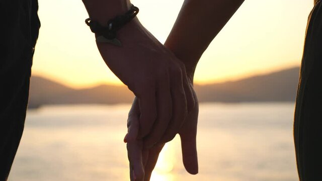 Male and female hands holding each other at sunset. Young couple standing on seashore and enjoying spending time together. Romantic moment between two lovers. Concept of loving or happiness. Slow mo