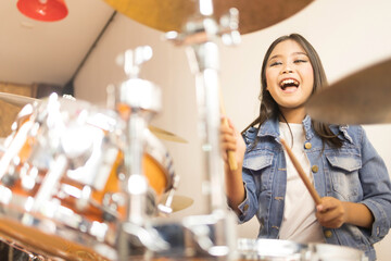 Fototapeta na wymiar A cute Asian elementary school girl with long hair and a denim jacket laughing happily while playing a drum in a music classroom.