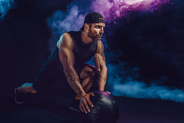 Aggressive bearded muscular sportsman is working out, push up with a medicine ball isolated on dark studio background with smoke.