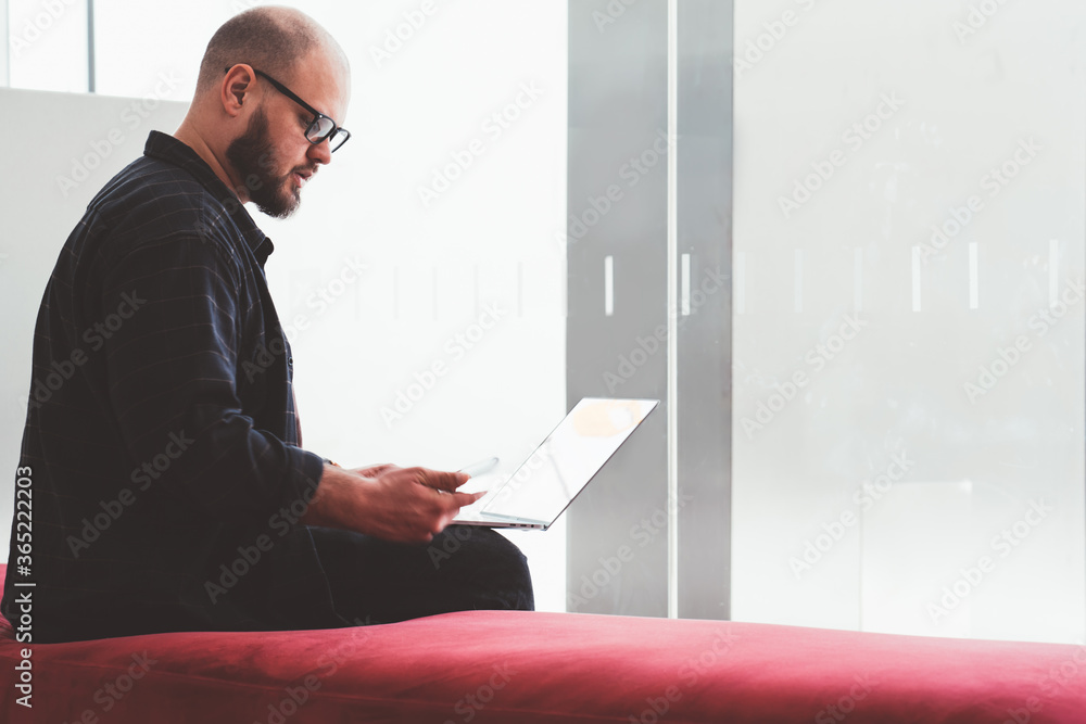 Poster pensive young man in eyeglasses reading notification on smartphone device during work at laptop comp