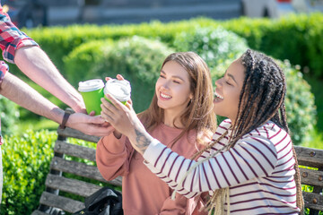 Two girls on bench taking coffee from hands of guy