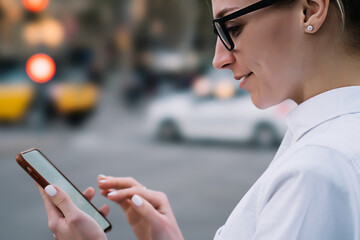 Fototapeta premium Cropped side view of smiling hipster blogger in eyeglasses holding modern smartphone in hand and reading incoming notification with good news.Positive young woman laughing while watching video online