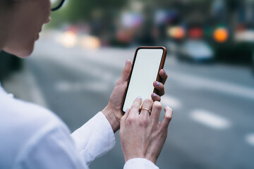 Cropped view of young woman holding smartphone device in hands and typing information on blank display of gadget on blurred background.Female fingers touching on blank screen of digital telephone