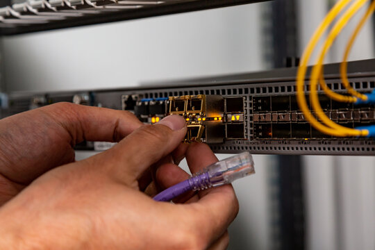 The electrician inserts the SFP module into the router and holds the patch cord. Hands in the frame. Small form-factor pluggable transceiver. Horizontal orientation. 