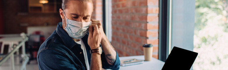 panoramic crop of businessman wearing medical mask near laptop with blank screen