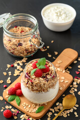 Parfait yogurt with granola and raspberries in a glass, black background, vertical orientation. Healthy breakfast concept.