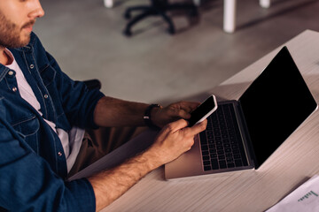 cropped view of businessman using smartphone near laptop on desk
