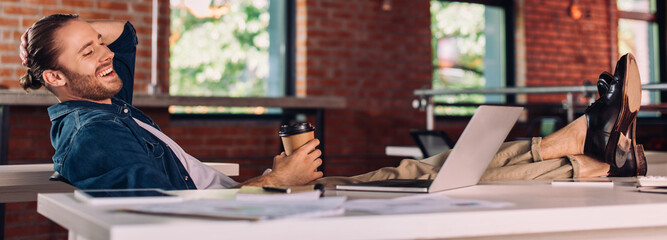 panoramic crop of cheerful businessman holding paper cup and looking at laptop in office