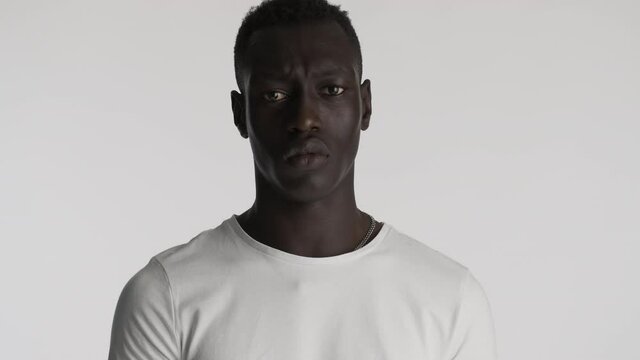 Young upset African American man in white t-shirt sadly looking in camera over gray background. Stressed expression