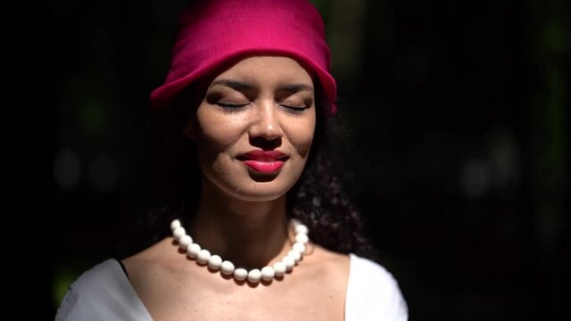 Close-up of a dark-haired curly-haired young smiling girl walking in a green summer Park. She wears large white beads and a lilac headscarf. She stands with her eyes closed and then opens them.