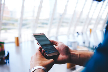 Cropped shot view of man's hands orders electronic ticket via mobile phone application during summer trip, young male reading text message on cell telephone while standing in waiting hall of airport