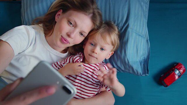 Top view Young beautiful mother with little baby lying in bed and making video call or photo. Woman blogger making selfies or video with child, happy parenting.