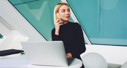 Young female economist having pleasant conversation on mobile phone while sitting at the table with...