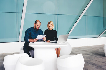 Male confident CEO reading monthly report on paper documents while sit with his secretary in office...