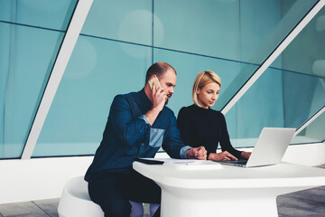 Young businessman talking on mobile phone with client while his secretary keyboarding on net-book,...