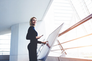 Businesswoman with cell telephone in hand touching big computer monitor while standing in modern office interior, young female looking away while verify data on mobile phone and interactive display