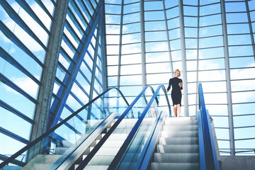 Young businesswoman is searching information on website via cell telephone,while is standing near escalator staircase in modern airport interior with contemporary building design Copy space background