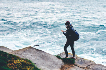 View from above of a young hipster girl with a rucksack on her back watching photos on cell telephone, stylish woman tourist using mobile phone while standing on a rock against blue sea with waves