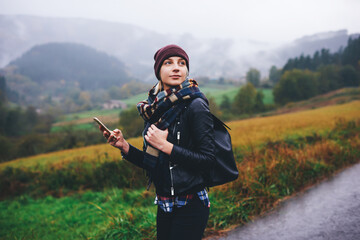 Trendy woman catches signal on her mobile phone while searching interesting places in mountains, beautiful female tourist with rucksack waiting for a text message on cell telephone during walking tour