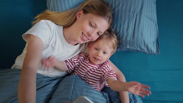 Young beautiful mother with little child in bed making video on phone, smiling and laughing, greeting followers, spending time together, happy family motherhood.