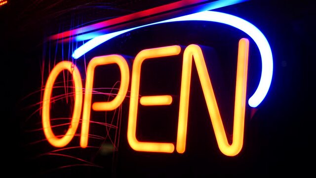 Open neon sign glowing in the dark. Vivid retro styled text at entrance on glass window. Colorful electric banner selective focus close up. Light bulbs radiance at night. Shiny illuminated lettering.