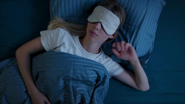 Young girl in bed in sleep mask, suffers from insomnia, tries to fall asleep, tosses, discontentedly removes sleep mask from eyes