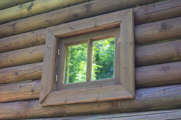 The wall of a dark brown log house made of coniferous logs with a window that reflects the forest,