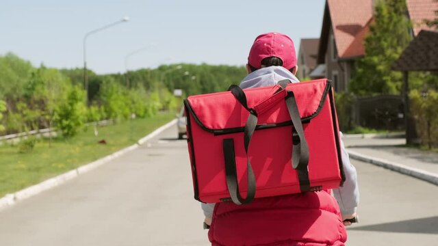 Tracking with tilt up of delivery man with insulated backpack riding bicycle along street in suburban neighborhood
