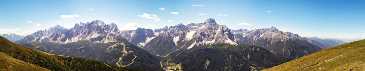 Panorama banner of the Alps mountains in Italy. Dolomites in summer with small snowfields on the peaks. Blue sky with small clouds, very green wooded trees.