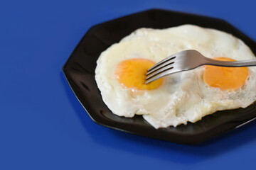 Close up of fork poking yolk of a delicious fried egg ready to eat on a blue background
