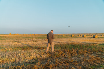 A young man stands on a wheat field with bales and controls the drone