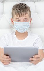Sick boy wearing medical protective mask uses tablet computer at home. Quarantine and coronavirus epidemic concept