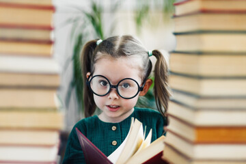 Cute smiling preschool age girl in round big glasses with red book in her hands between two books piles
